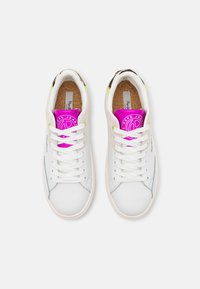 Pár bílých tenisek s béžovými akcenty, veselými růžovými štítky na jazyku, bílými tkaničkami a korkovými vložkami, pohled zeshora na bílém pozadí.