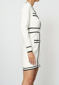 Robe en tricot blanche à manches longues, avec des rayures texturées, des accents noirs et des détails en boutons dorés. Elle présente une silhouette ajustée et des poches sur les côtés.