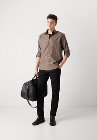 Bruine overhemd met knopen, zwarte broek, slanke zwarte leren laarzen en een grote zwarte leren tas, geposeerd tegen een neutrale achtergrond.