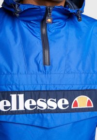 Klart blå jacka med en svart dragkedja och ett stort marinblått band som visar vit "ellesse"-text och en röd, orange och gul logotyp.