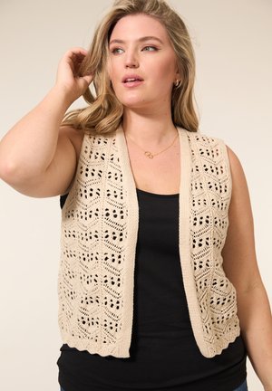 Leren haakbrei mouwloze vest in beige met een open patroon en geschulpte randen, gedragen over een zwart tanktop. Eenvoudig, ontspannen ontwerp.