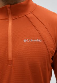 Primer plano de una persona que lleva una chaqueta con cremallera naranja de Columbia con tela texturizada y detalles visibles de costuras.