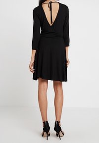 Svart omlottklänning med långa ärmar, djup V-formad rygg och flödande kjol. Har en knytning vid halsen och är i kombination med högklackade skor.