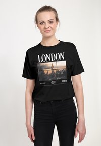 Černé tričko s potiskem, na kterém je text "LONDÝN", fotografie Big Benu a čárový kód. Látka vypadá hladce a má volný střih.