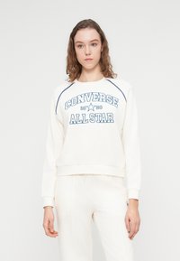 Sweat-shirt crème avec texte bleu "Converse All Star", poignets et ourlet côtelés, et manches raglan ; assorti à un pantalon crème assorti.