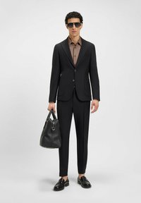 Zwarte pak met een getailleerde blazer en broek, een bruine blouse eronder, gecombineerd met zwarte instappers en een gestructureerde zwarte handtas.