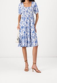 Robe à imprimé floral bleu avec un décolleté carré, des manches bouffantes et une jupe évasée. Portée avec des talons beige à fines lanières et un petit sac à main blanc.