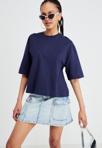 Marineblå bomulds T-shirt med korte ærmer, parret med en lyseblå denimskørt med lommer. Modellen bærer rosa solbriller og hoops øreringe.