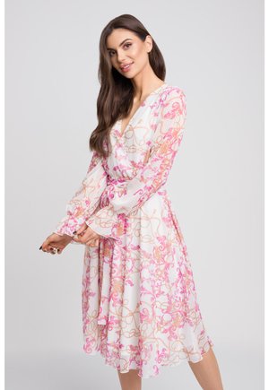 Kwiatowa sukienka w stylu wrap z przejrzystego białego materiału, zdobiona różowymi i brzoskwiniowymi falami oraz łańcuchami. Długie rękawy z falbaniastymi mankietami i zwiewna spódnica.