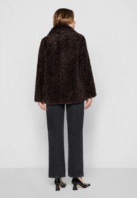 Veste en fausse fourrure marron avec une coupe ample, un col montant et des manches longues. Portée sur un pantalon à larges jambes noir et des chaussures à talons noires.