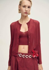 Roter, transparenter Cardigan mit Knöpfen über einem passenden, cropped Top. Der Rock hat eine silberne, runde Gürtelakzentuierung und ein rotes Blumenmotiv.