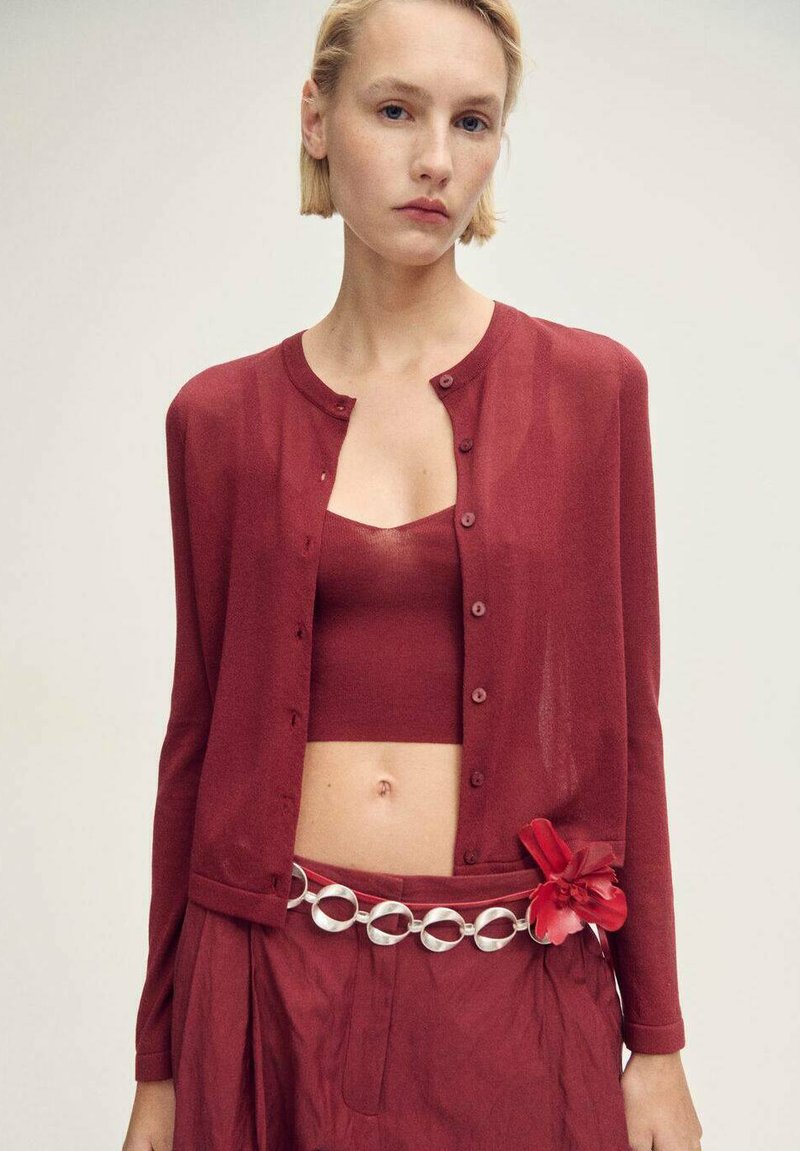 Roter, transparenter Cardigan mit Knöpfen über einem passenden, cropped Top. Der Rock hat eine silberne, runde Gürtelakzentuierung und ein rotes Blumenmotiv.