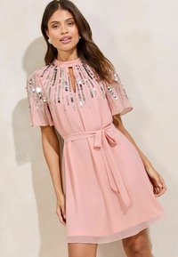 Kurzes pinkes Kleid mit einem Schlüssel-ausschnitt, kurzen Ärmeln und Paillettenverzierungen an den Schultern. Mit einem gebundenen Gürtel in der Taille.