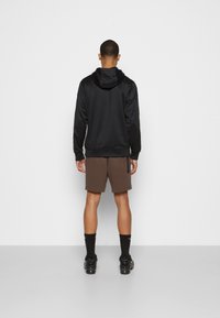 Svart hoodie med en slät textur, tillsammans med bruna shorts. Mörka strumpor och svarta skor fulländar outfiten. Sedd bakifrån mot en neutral bakgrund.