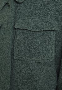 Grøn fleecejakke med en tekstureret overflade og en klaplomme. Stoffet er blødt og tykt, hvilket giver isolering og komfort.