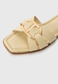 Beige leather slide sandal with a square toe, featuring a prominent gold buckle and crossed strap design, smooth texture and minimal stitching.