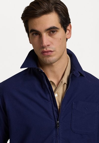 Chaqueta de punto color navy con un alto cuello, bolsillo lateral y una textura suave, superpuesta sobre una camisa beige con cuello.