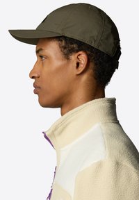 Olive green cap with a curved brim, featuring stitched details. Worn alongside a cream fleece jacket with purple accents and a zip closure.