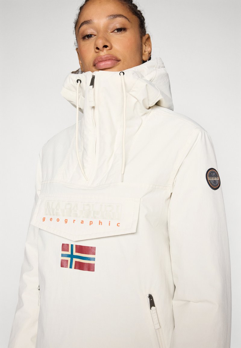 Primer plano de una persona que lleva una chaqueta blanca de Napapijri con un alto cuello, un bolsillo frontal y un parche de la bandera noruega debajo de la solapa del bolsillo.