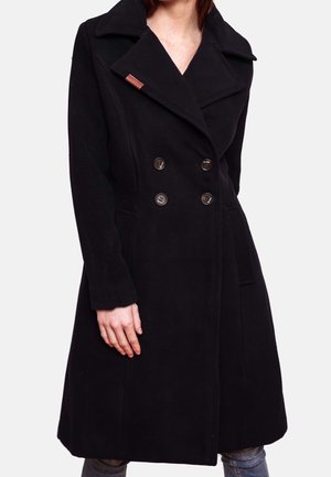 Frau trägt einen knielangen schwarzen doppelreihigen Mantel mit großen Revers, Knopfdetails und seitlichen Taschen, steht mit einer sichtbaren Hand.