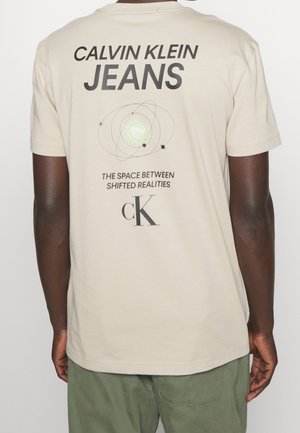 Homme portant un t-shirt beige avec le texte noir "Calvin Klein Jeans", un graphisme orbital et l'inscription "L'espace entre les réalités décalées" au dos.