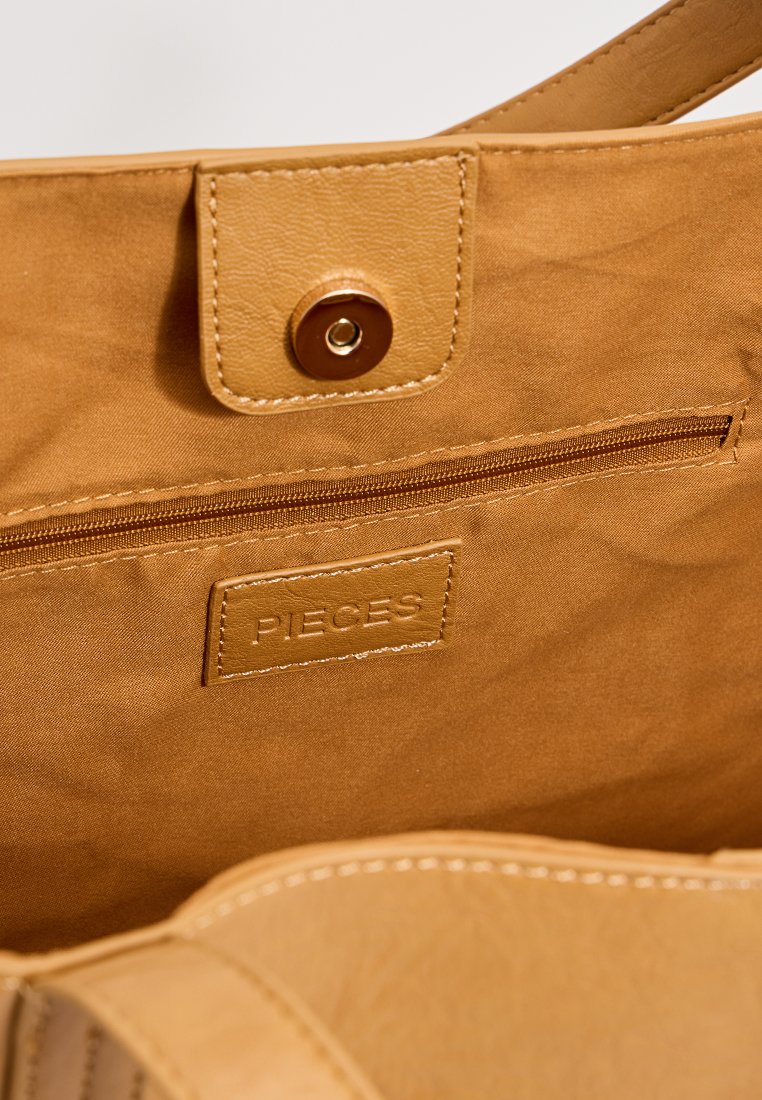 Intérieur d'un sac fourre-tout beige avec une poche zippée, marqué "PIECES", présentant des accents en cuir lisse et une doublure en tissu doux et texturé.