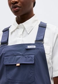 Άτομο με σκουρόχρωμο δέρμα που φοράει λευκό πουκάμισο με γιακά κάτω από σκούρο μπλε παντελονοφόρεμα Columbia με ρυθμιζόμενες τιράντες και μπροστινή τσέπη.