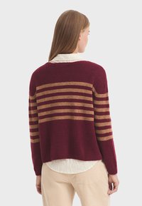 Maglione bordeaux con strisce orizzontali color sabbia, texture a maglia, collo rotondo e vestibilità comoda. Indossato sopra una camicia chiara con colletto.