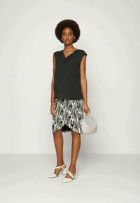 Top preto sem mangas com decote rendado, combinado com uma saia envolvente preta e branca com padrões, sapatos brancos de salto e uma pequena bolsa cinza.