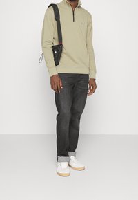 Lehký olivově zelený pulovr na zip, černá taška s popruhem, tmavé džíny, bílé tenisky s béžovými podrážkami. Minimalistický design, hladká textura, volný střih.