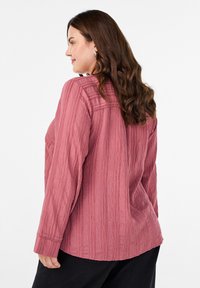 Žena s dlhými vlnitými hnedými vlasmi, oblečená v štruktúrovanej dymovo ružovej blúzke s dlhým rukávom a čiernych nohaviciach, otočená mierne na stranu, proti bielej stene.