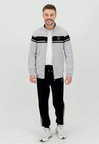Gray zip-up jacket with black stripes and gold logo, paired with black joggers featuring white side stripes and pockets. White sneakers complete the look.