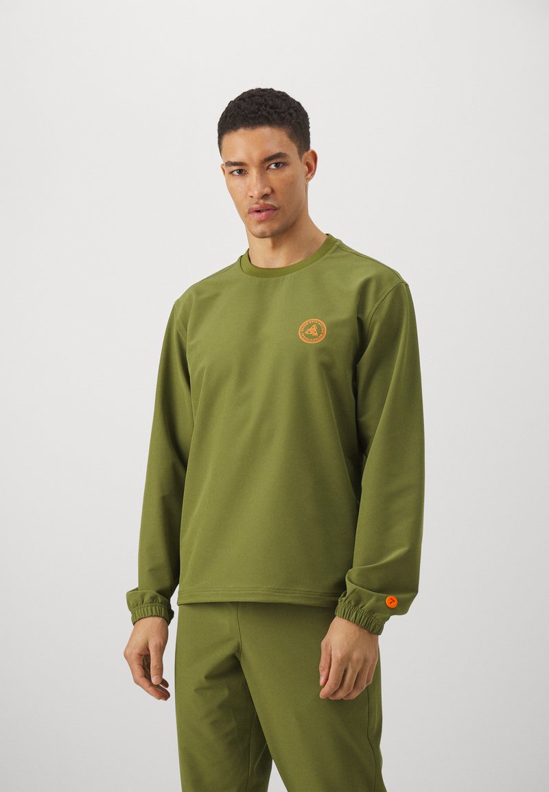 Olive green long-sleeve shirt with an elastic cuff, featuring a circular orange logo on the chest. Soft, matte fabric finish.