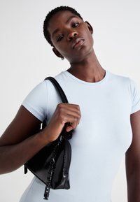 T-shirt aderente di colore azzurro chiaro con maniche corte, caratterizzato da un collo rotondo. Borsa nera con finitura testurizzata e dettagli con zip tenuta in mano.