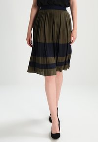 Jupe plissée longueur genou avec des rayures alternées vert olive et bleu marine. La ceinture est élastique, créant une silhouette lisse.