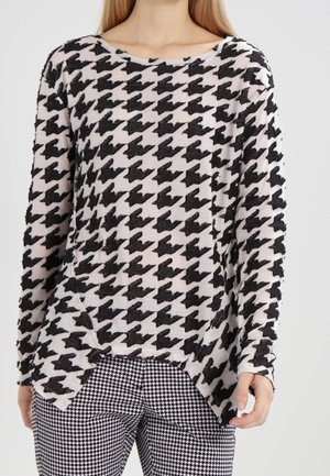 Pitkähihaine neule pehmeästä kankaan, jossa on musta ja kerma houndstooth-kuvio, pyöreä kaula-aukko ja sivuhalkiot hemuksessa.
