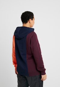 Sudadera con capucha que presenta un diseño a bloques de color en naranja, azul marino y burdeos. Fabricada con una tela suave y con puños y dobladillo de canalé.