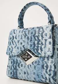 Borsa in denim con tonalità blu, caratterizzata da una chiusura a patta, una placca del logo prominente e un motivo in rilievo con lettere su tutta la superficie.