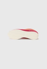 Chaussures de sport avec une tige rouge et orange, semelle en caoutchouc blanc présentant un motif en zigzag. Texture douce et forme arrondie.