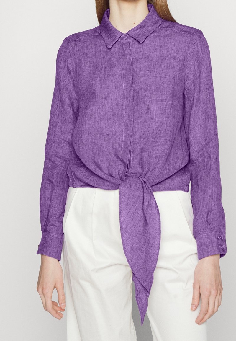 Femme portant une chemise violette à manches longues boutonnée et nouée à la taille, associée à un pantalon blanc taille haute, sur un fond uni.