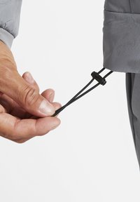 Gray puffer jacket cuff with a black adjustable drawstring. A hand grips the cord, showcasing material texture and cinching mechanism.