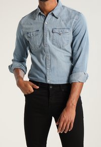 Ljusblå denimskjorta med uppvikt lång ärm, två bröstfickor och knappstängning; matchad med åtsittande svarta jeans.