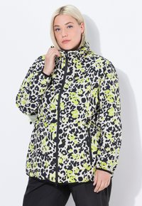 Donna bionda che indossa una giacca a zip nera, bianca e giallo neon con motivo e tasche, mentre sistema il colletto, su uno sfondo bianco.