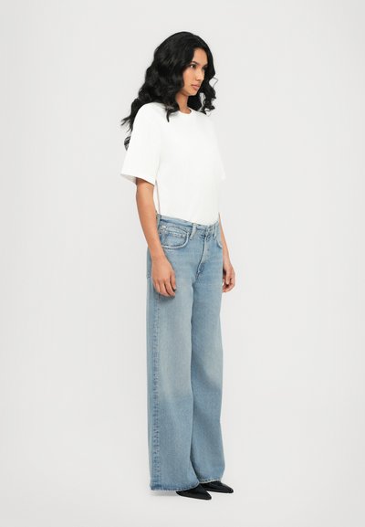 T-shirt blanc à manches courtes associé à un jean large bleu clair. Le modèle se tient de côté, portant des chaussures noires à talons pointus et ayant de longs cheveux ondulés.