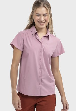 Junge Frau lächelnd, trägt ein kurzärmliges, rosa Knopfhemd und rostfarbene Hosen, steht vor einem schlichten hellen Hintergrund.