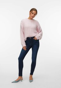Femme portant un pull rose en maille torsadée, un jean skinny bleu foncé et des escarpins à bride arrière bleu clair, debout avec une main dans la poche.