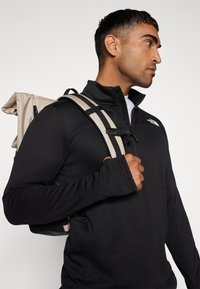 Veste zippée noire en tissu texturé, associée à un sac à dos beige avec des bretelles rayées. Matériel de sangle ajustable visible.