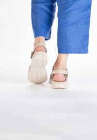 Beige sandals with adjustable strap, rubber sole featuring grooves for traction, worn with loose blue linen pants.
