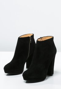 Botines de ante negros con un alto tacón cuadrado, punta redonda y cierre de cremallera lateral. Textura suave con un diseño minimalista.