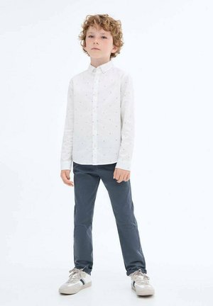Chemise blanche à manches longues avec des motifs discrets, associée à un pantalon bleu marine. L'enfant porte des baskets de couleur claire avec des accents.